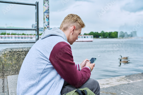 Fotomurale Serious guy college student using smart phone sitting on city river embankment
