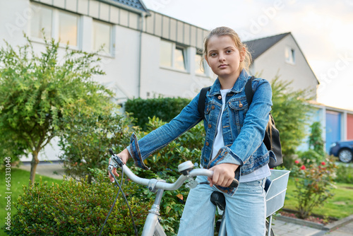 Schoolgirl child 10, 11 years old with backpack on bicycle on street near house