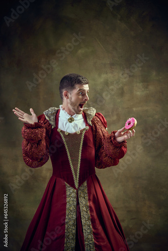 Canvas Print Artistic young man, royal person in female dress looking on pink donut against dark green background