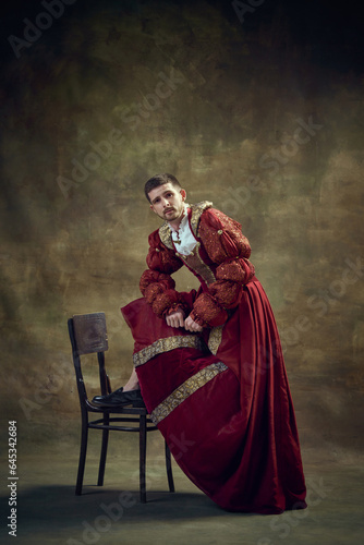 Young medieval prince, royal person leaning on chair, wearing female dress on dark green background. Renaissance style. Historical retrospectives, fashion, provoking projects, gender fluidity concept