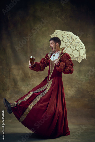 Joyful man, medieval prince walking in female dress with umbrella and drinking beer against vintage background. Concept of historical retrospectives, fashion, provoking projects, gender fluidity