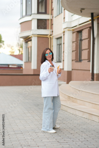 A young beautiful fashionable woman is walking near a modern building and smile with glass of coffee and yellow book..