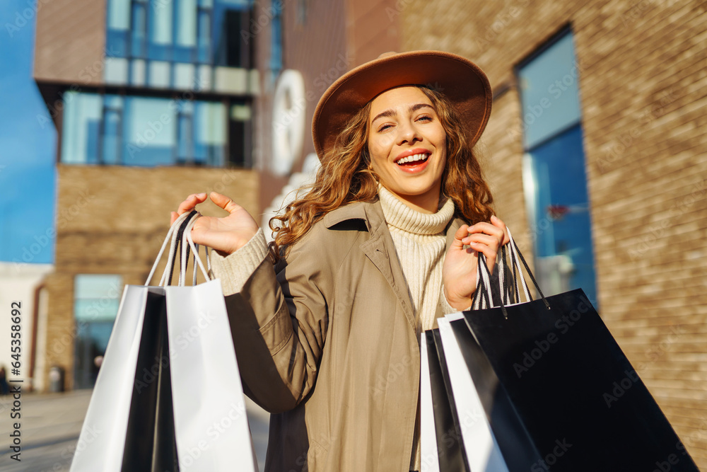 Portrait of a stylish woman in a hat after shopping near the store. Curly-haired woman with shopping bags walks through a sunny city. Concept of sales, shopping. Portrait of a stylish woman in a hat after shopping near the store. Curly-haired woman with shopping bags walks through a sunny city. Concept of sales, shopping.