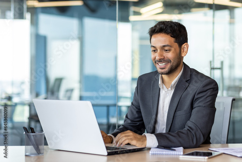 Portrait of young arab businessman inside office, man typing on laptop smiling, boss happy with results of financial success at workplace. © Liubomir