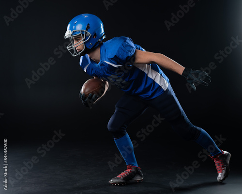 Full length portrait of a man in a blue american football uniform against a black background. Sportsman in a helmet with a ball. 