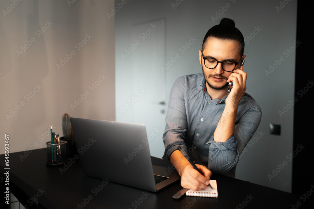 Portrait of young serious businessman with hair bun, working home at laptop, makes notes in notepad, talking on smartphone with clients, wearing eyeglasses and shirt. Background of grey wall. Portrait of young serious businessman with hair bun, working home at laptop, makes notes in notepad, talking on smartphone with clients, wearing eyeglasses and shirt. Background of grey wall.