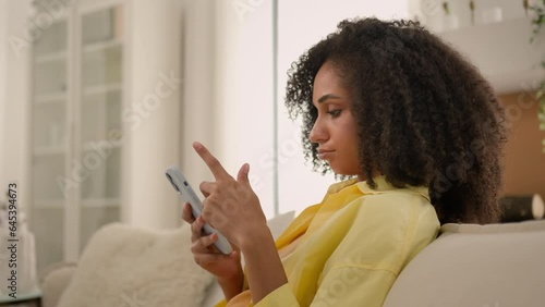 Bored African American ethnic woman gadget addict girl chatting online smartphone overuse social media addiction lazy sad female homeowner on cozy sofa at home mobile phone scrolling news on cellphone