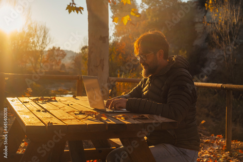 One man alone working on laptop in outdoor sitting on a wooden table at the park during a golden autumn amazing sunset. People and small business or digital nomad technology roaming lifestyle online