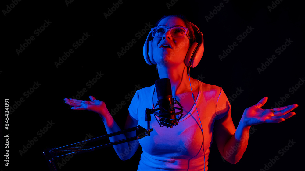 Caucasian woman in glasses and headphones sings into a microphone in neon light on a black background. An emotional girl is recording a song in a recording studio Caucasian woman in glasses and headphones sings into a microphone in neon light on a black background. An emotional girl is recording a song in a recording studio