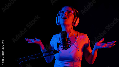 Caucasian woman in glasses and headphones sings into a microphone in neon light on a black background. An emotional girl is recording a song in a recording studio