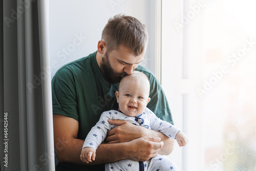 Photographie Father hugs baby boy, young man kisses his son on head in living room