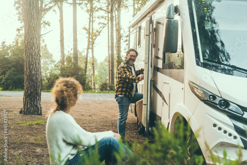 Adult couple enjoy outdoor leisure activity together outside a modern camper van motorhome. Travel people vacation lifestyle. Parking in a middle of nature with forest and trees in background. Freedom
