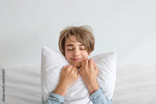 Handsome smiling teenager boy with eyes closed embracing white pillow, sitting on bed, white wall background. Teenager healthy lifestyle, sleep routine, rest, good sleep time concept