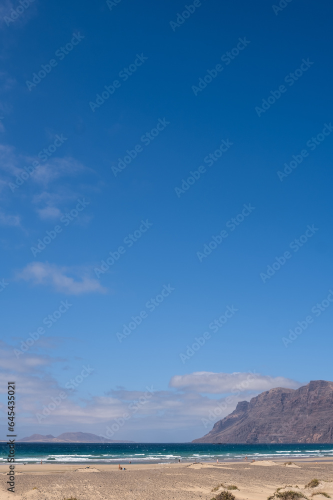 Fototapeta premium White sand beach. Famara beach. Turquoise water, island of La Graciosa and cliffs in the background. Sky with big white clouds. Caleta de Famara.Lanzarote, Canary Islands, Spain.
