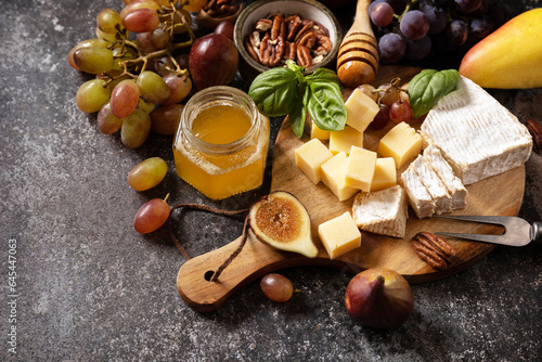 Brie cheese, fresh figs, grapes, cheese cubes, pears, pecan nuts and honey on cutting board on dark stone background. Cheese plate, still life of seasonal wine snacks.