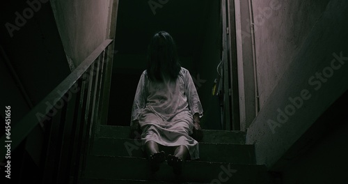 Horror scene of a mysterious Scary Asian ghost woman creepy have hair covering the face sitting on staircase at abandoned house with background dark scene movie at night, festival Halloween concept