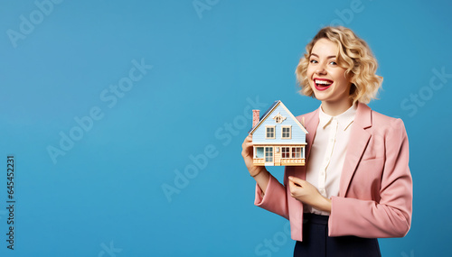 Portrait of young woman with happy positive smile hands holding house miniature isolated on blue background, copy space, moving to a new house, buying real estate