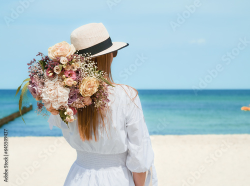 beautiful young woman with a bouquet of flowers on the beach. pretty woman in white dress and sun hat. Vacation and freedom concept.