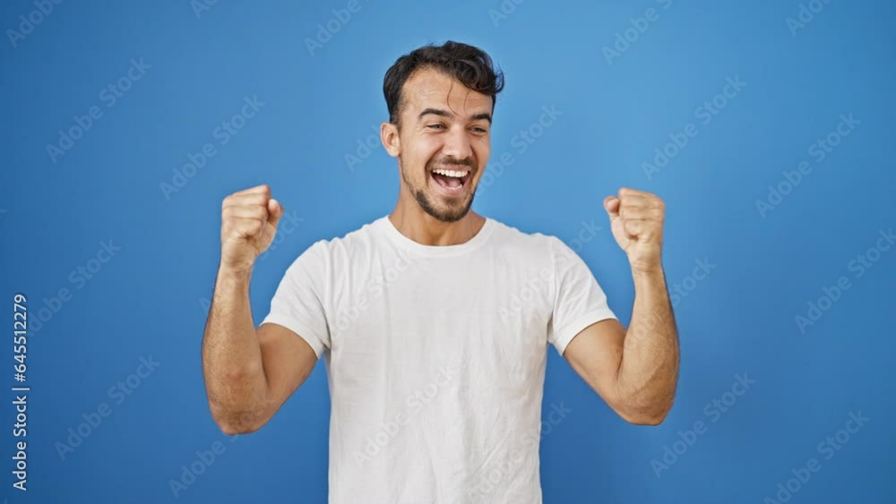 Young hispanic man smiling confident standing with winner gesture over isolated blue background Young hispanic man smiling confident standing with winner gesture over isolated blue background