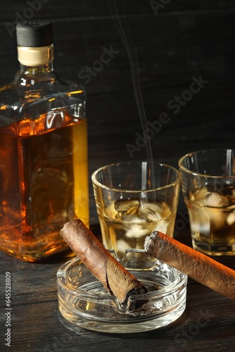 Cigars, ashtray and whiskey with ice cubes on black wooden table