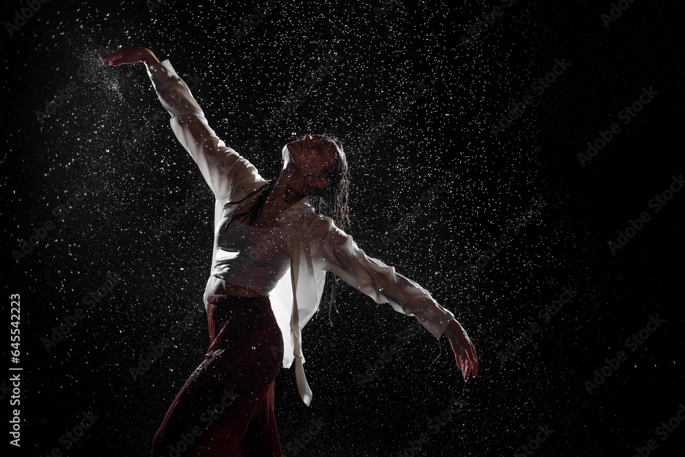 Fototapeta premium Dancing in the rain. A young woman in wet clothes dancing among water droplets, photo in the aqua studio. beautiful silhouette, model in a white shirt