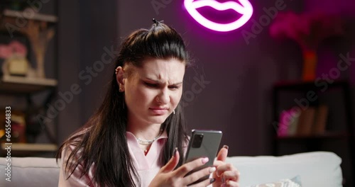 Woman scrolls through feed on social networks from smartphone. Woman frowns, looking at phone screen with disgruntled incredulous face. Feelings of jealousy, abhorrence, disgust. Unpleasant nasty news photo