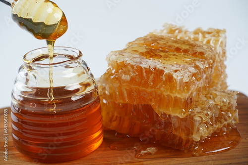 Honey jar and stick with liquid honey flowing on honeycomb pieces in a puddle.