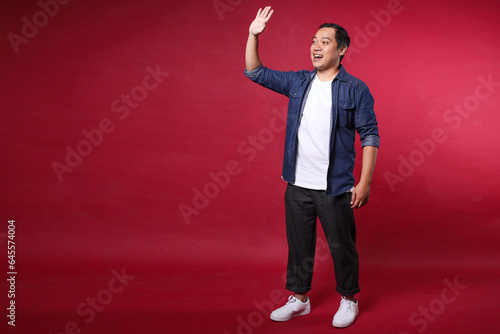 Full body portrait of casual asian man with say Hi gesture and smiling away  on red background © Gatot