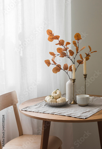 Autumn home interior in the living room - around wooden table, a chair, a bouquet of autumn leaves in a vase, decorative pumpkins. Cozy home