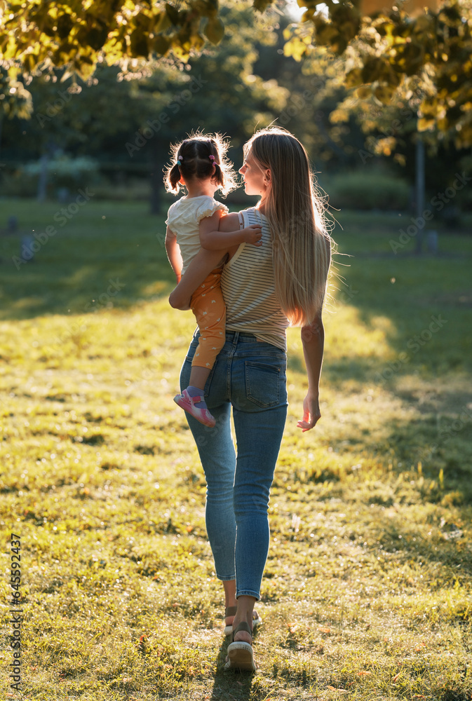 Fototapeta premium young mother with little daughter. Mom walks in the park with her daughter and holds her in her arms. view from the back