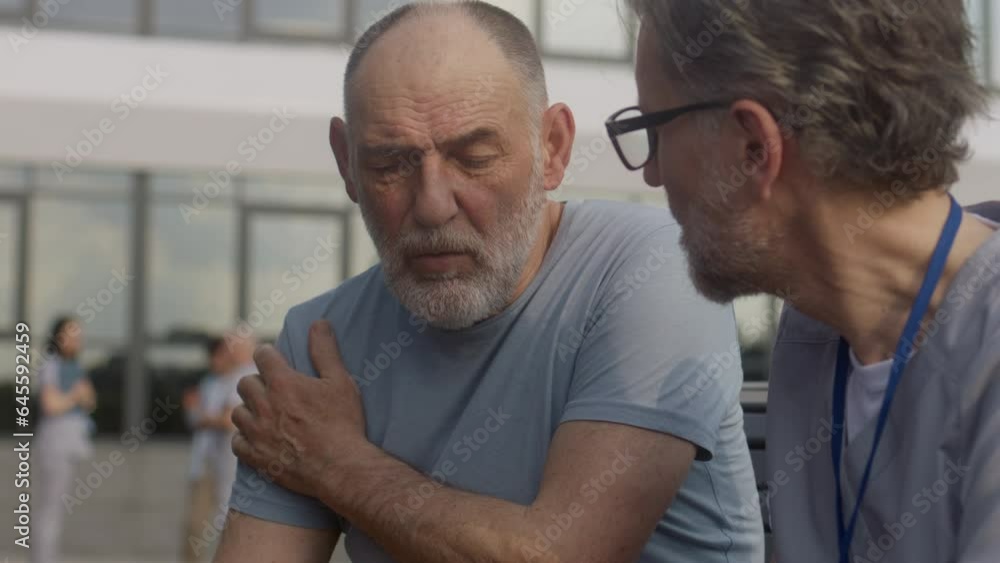 Health care specialist sits with aged patient on bench and discusses his treatment. Elderly man complains of shoulders and heart pains to mature doctor. Professional in medical uniform works outdoor. Health care specialist sits with aged patient on bench and discusses his treatment. Elderly man complains of shoulders and heart pains to mature doctor. Professional in medical uniform works outdoor.