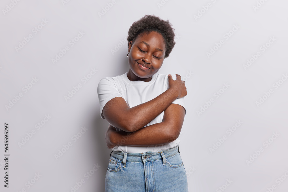 Love yourself. Dark skinned plump woman hugs herself recalls something in mind wears casual t shirt and jeans isolated over white background smiles carefree and happy feeling comfortable and fulfilled - obrazy, fototapety, plakaty 