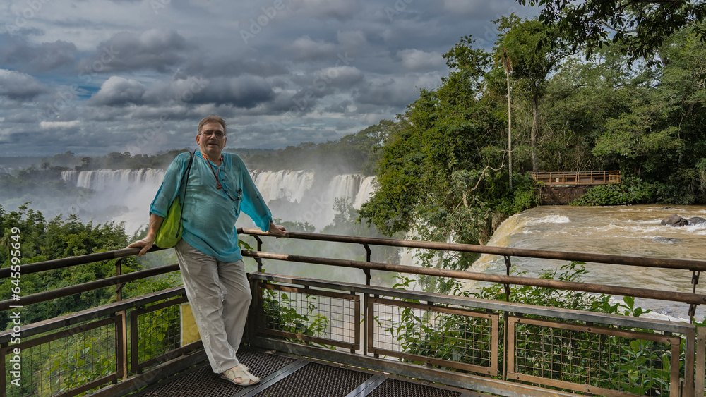 The man is standing on the observation deck at the waterfalls, leaning on the railing, smiling. Behind- are the picturesque Iguazu Falls, shrouded in spray and fog. Nearby is lush tropical vegetation 