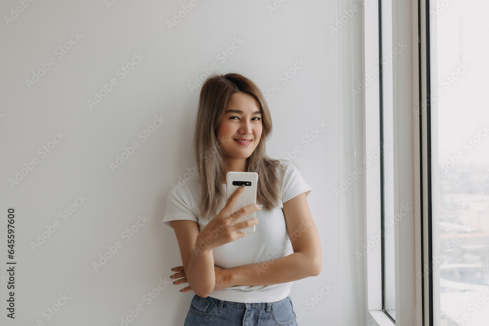Asian Thai woman holding cellphone and looking at the camera, Smiling and standing next to the window at room apartment. Asian Thai woman holding cellphone and looking at the camera, Smiling and standing next to the window at room apartment.