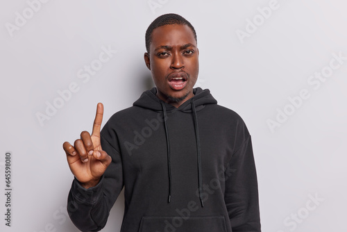 Horizontal shot of serious dark skinned adult man points index finger having hearing problem listening to something says attention please wears casual sweatshirt isolated over white background