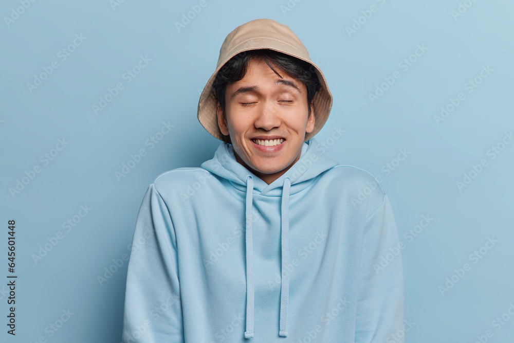 Horizontal shot of handsome cheerful Asian man keeps eyes closed giggles positively wears panama and casual hoodie poses against blue background recalls something pleasant in mind. Emotions concept Horizontal shot of handsome cheerful Asian man keeps eyes closed giggles positively wears panama and casual hoodie poses against blue background recalls something pleasant in mind. Emotions concept