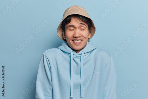 Horizontal shot of handsome cheerful Asian man keeps eyes closed giggles positively wears panama and casual hoodie poses against blue background recalls something pleasant in mind. Emotions concept photo