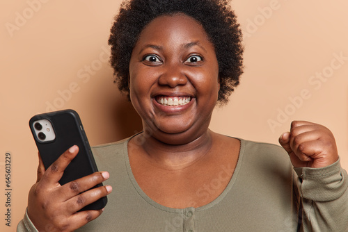 Positive overjoyed dark skinned obese woman smiles happily and clenches fist holds mobile phone reads good news rejoices nice relevation dressed in casual jumper isolated over beige background