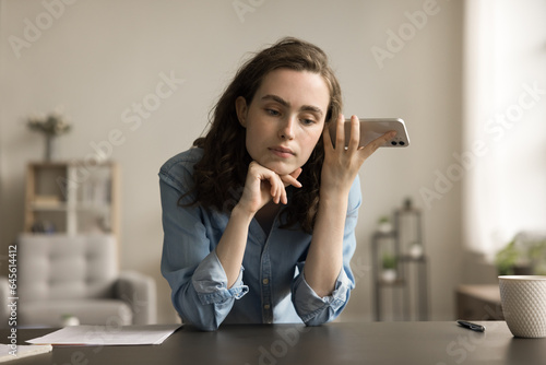 Serious freelancer employee girl holding mobile phone at head, listening to sound, voice message, audio file, leaning hands on home workplace table, thinking, touching chin