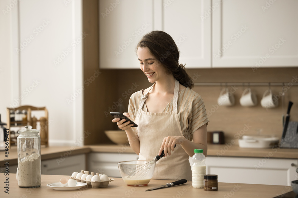 Happy beautiful young chef woman using smartphone over table with bakery food ingredients, preparing breakfast, taking picture, photo, cooking pancakes, omelet, reading online recipe on Internet Happy beautiful young chef woman using smartphone over table with bakery food ingredients, preparing breakfast, taking picture, photo, cooking pancakes, omelet, reading online recipe on Internet