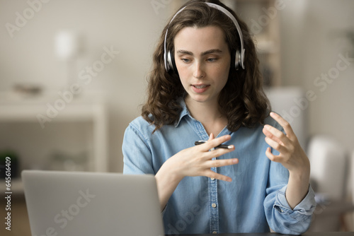 Serious gen Z student girl in headphones talking on video call at laptop, sitting at home office workplace. Online speaker streaming on Internet, giving workshop, webinar, speaking at conference