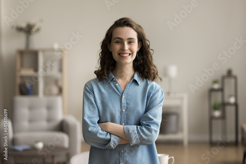 Cheerful confident gen Z young adult girl home head shot. Happy beautiful female student, freelance employee in casual posing in apartment with hands folded, looking at camera with toothy smile
