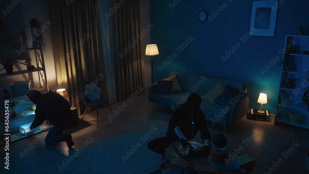 Shot of two burglars going around the apartment, looking for valuables, jewelry. The men are using flashlights to navigate in space. Night time, low, muffled light.