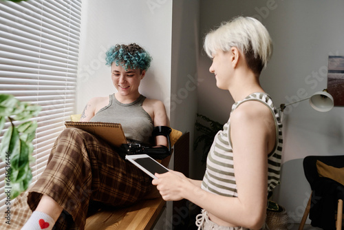Young girl sitting on windowsill and drawing in her notebook while her girlfriend using tablet pc