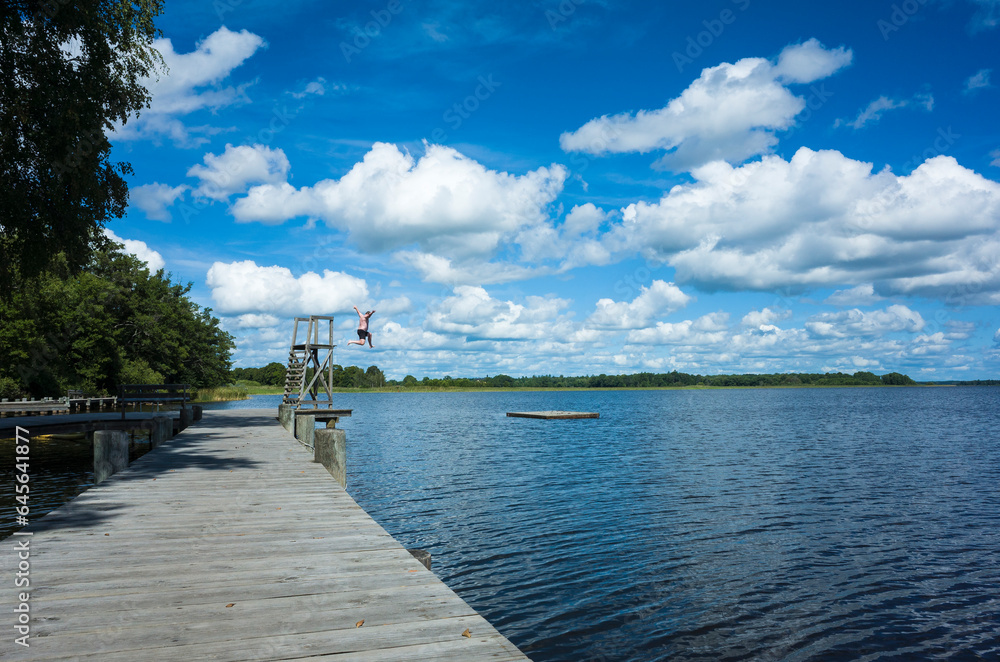 Summer outdoor activities in Sweden, Wooden bridge and jumping tower on Freden bay in lake Mälaren, Strömsholm, Man jumps in water, sunny day blue sky fluffy white clouds, Scandinavian landscape