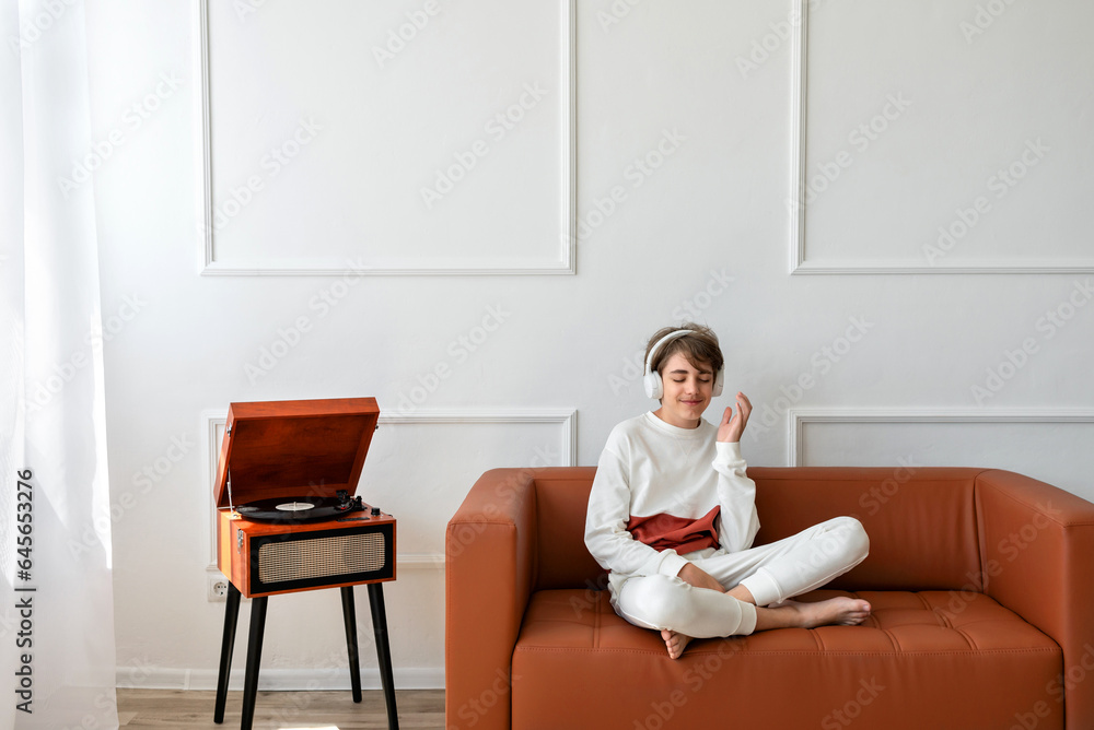 Happy smiling teenage boy sitting on sofa wearing earphones and listening music near turntable with vinyl record Happy smiling teenage boy sitting on sofa wearing earphones and listening music near turntable with vinyl record