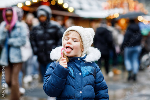 Little cute preschool girl with candy cane from a sweets stand on Christmas market. Happy child on traditional family market in Germany. Preschooler in colorful winter clothes during snowfall