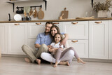 Positive attractive young couple of parents holding cheerful toddler kid in arms, sitting on heating floor in home kitchen, leaning on new furniture, looking away, laughing. Happy family portrait
