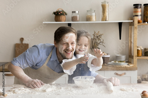 Joyful toddler kid and daddy enjoying funny baking activity with happy floury faces, throwing flour at kitchen table, hugging, smiling, laughing. Dad and daughter preparing bakery food
