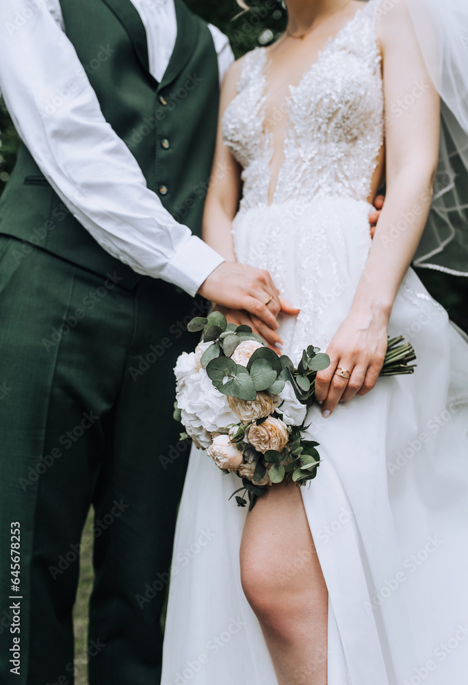 The groom in a green suit and the bride in a white lace dress hold hands with golden rings on their fingers and a bouquet of flowers at the ceremony. Wedding photography, close-up portrait.